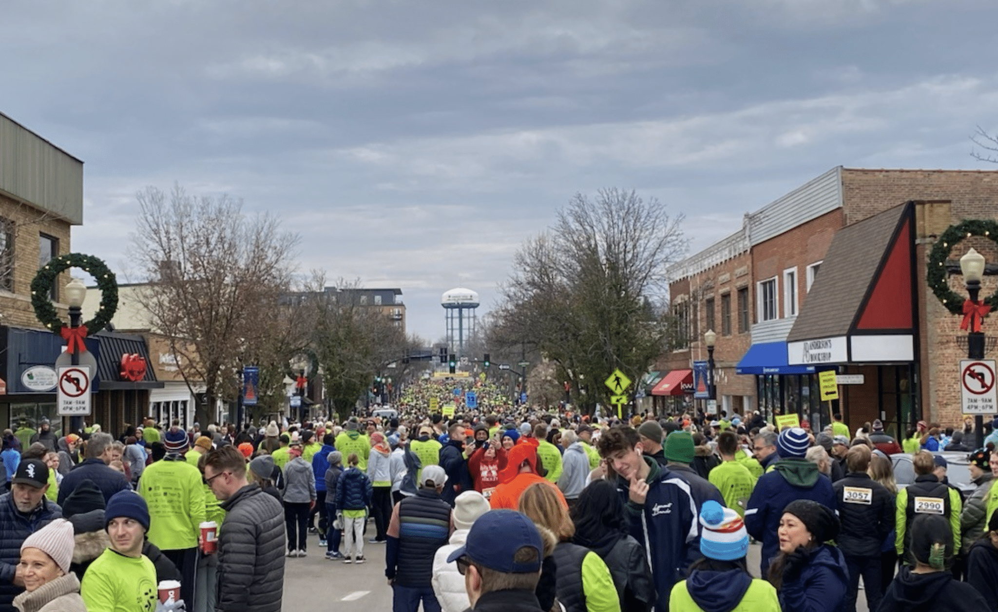 Crowd of participants in bright green shirts at a festive event in Downers Grove, with shops decorated for the holidays and a water tower in the background.